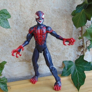 May include: A detailed action figure of a character in a black and red suit, with a menacing expression. The figure has a spider-like design on the chest and head, with red accents on the arms and feet. The figure is posed with arms outstretched.