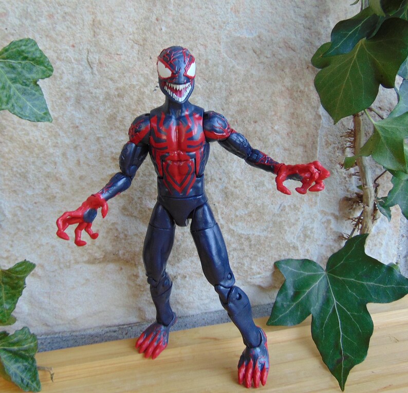May include: A detailed action figure of a character in a black and red suit, with a menacing expression. The figure has a spider-like design on the chest and head, with red accents on the arms and feet. The figure is posed with arms outstretched.