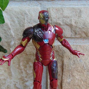 May include: An Iron Man action figure featuring a red and gold metallic suit with a glowing blue arc reactor on the chest. The figure has a detailed helmet with gold faceplate accents and articulated limbs posed with arms extended outward.