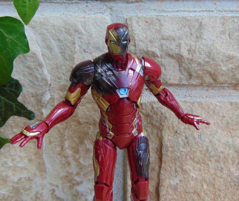May include: An Iron Man action figure featuring a red and gold metallic suit with a glowing blue arc reactor on the chest. The figure has a detailed helmet with gold faceplate accents and articulated limbs posed with arms extended outward.