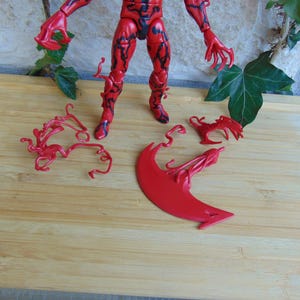 May include: A red action figure with black detailing, posed on a wooden surface. Several red, stylized accessories are placed around the figure. The figure has a muscular build and claw-like hands. The overall theme is a superhero or villain.