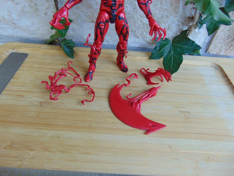 May include: A red action figure with black detailing, posed on a wooden surface. Several red, stylized accessories are placed around the figure. The figure has a muscular build and claw-like hands. The overall theme is a superhero or villain.