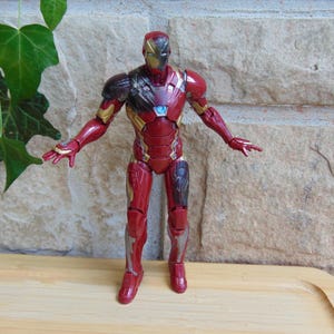May include: An Iron Man action figure featuring a metallic red and gold suit with a glowing light blue arc reactor on the chest. The figure stands in an upright position with arms extended outward, showcasing detailed armor plating and joints.