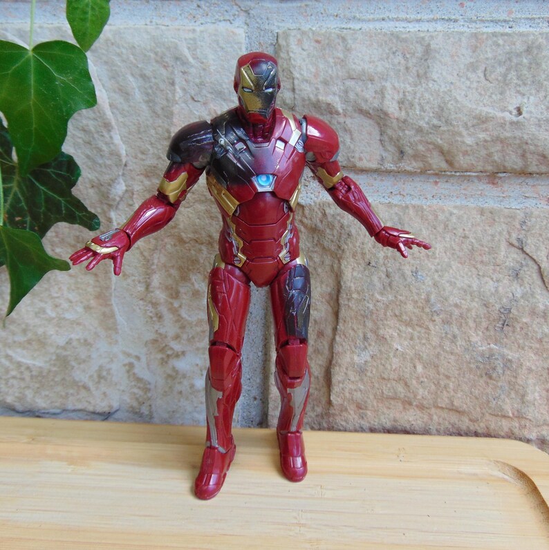 May include: An Iron Man action figure featuring a metallic red and gold suit with a glowing light blue arc reactor on the chest. The figure stands in an upright position with arms extended outward, showcasing detailed armor plating and joints.