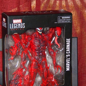 May include: A Marvel Legends action figure of Marvel's Carnage, a red and black character with a long tongue, in its original packaging. The box is black with the Marvel Legends Series logo and the text "MARVEL'S CARNAGE".