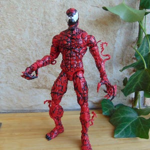 May include: A red Carnage action figure with black vein-like patterns. The figure has a white face with black details and sharp claws. The figure is posed with arms outstretched. The figure is a collectible.