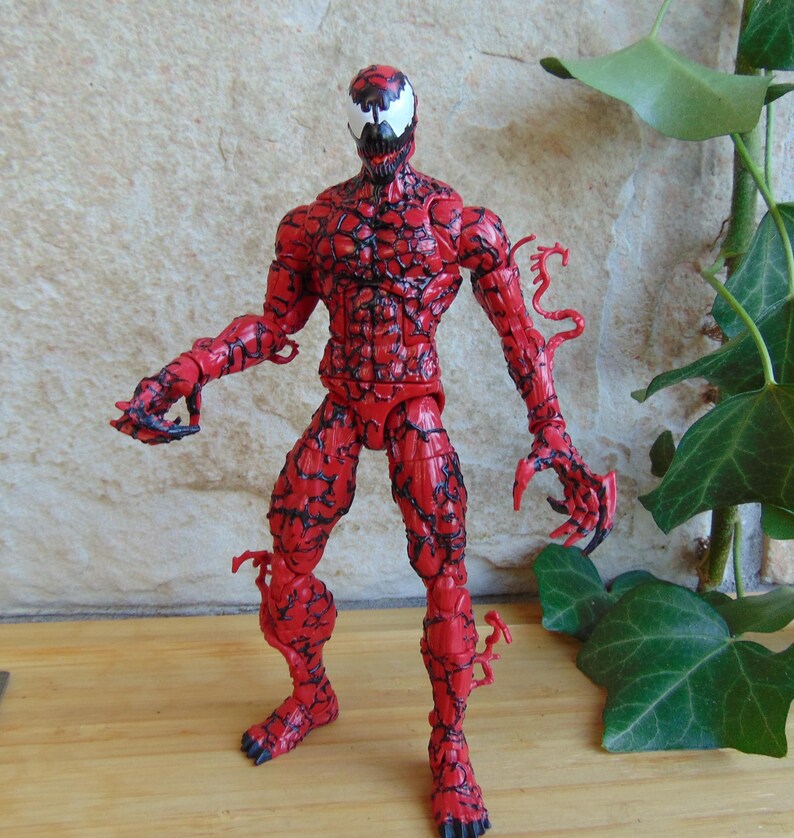 May include: A red Carnage action figure with black vein-like patterns. The figure has a white face with black details and sharp claws. The figure is posed with arms outstretched. The figure is a collectible.