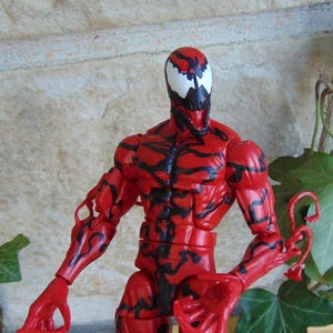 May include: A red action figure of the character Carnage, with black vein-like patterns across its body. The figure has a white face with black accents and a menacing open mouth. The figure is posed with arms outstretched.
