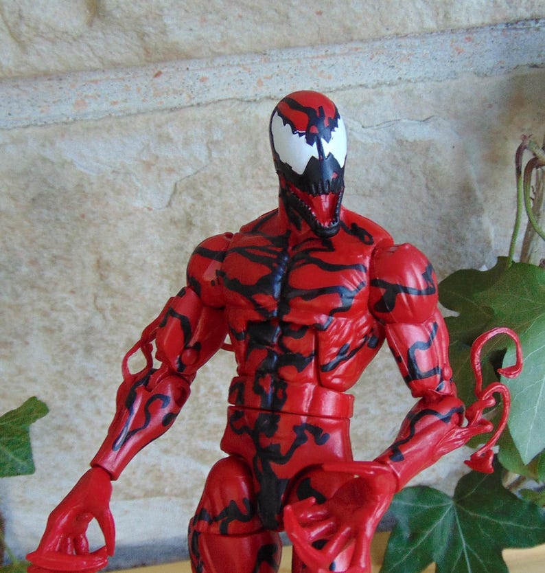 May include: A red action figure of the character Carnage, with black vein-like patterns across its body. The figure has a white face with black accents and a menacing open mouth. The figure is posed with arms outstretched.