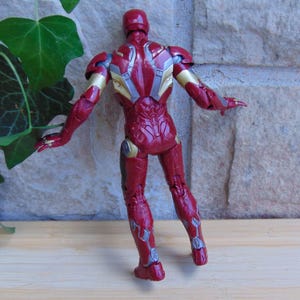 May include: A red and gold action figure of a humanoid character in armor, viewed from the back. The figure features intricate mechanical detailing across the torso, arms, and legs, with a glossy finish and articulated joints throughout the body.