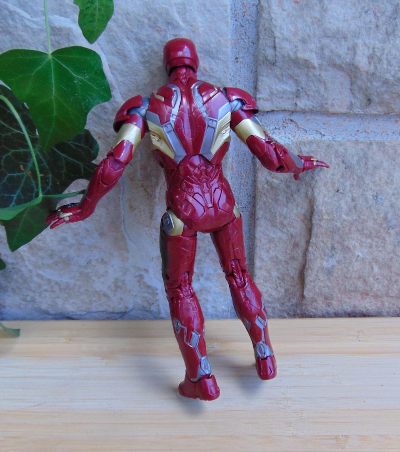 May include: A red and gold action figure of a humanoid character in armor, viewed from the back. The figure features intricate mechanical detailing across the torso, arms, and legs, with a glossy finish and articulated joints throughout the body.