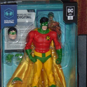 Puede incluir: Figura de acción de Robin de McFarlane Toys, empaquetada en una vitrina transparente. La figura es roja, verde y amarilla, con cabeza y capa extraíbles. El embalaje incluye el texto "McFarlane Toys Digital".