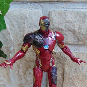 May include: An Iron Man action figure featuring a red and gold armored suit with a metallic finish. The figure has a glowing blue circular arc reactor on the chest, a detailed helmet, and articulated limbs posed with arms extended outward.