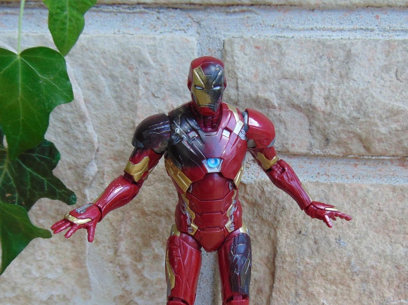 May include: An Iron Man action figure featuring a red and gold armored suit with a metallic finish. The figure has a glowing blue circular arc reactor on the chest, a detailed helmet, and articulated limbs posed with arms extended outward.