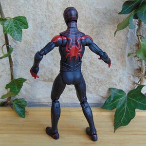 May include: A black and red action figure of a superhero, viewed from the back. The figure has a black suit with red accents on the shoulders, head, and a spider emblem on the back. The figure is posed with arms outstretched.