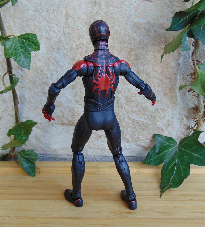 May include: A black and red action figure of a superhero, viewed from the back. The figure has a black suit with red accents on the shoulders, head, and a spider emblem on the back. The figure is posed with arms outstretched.