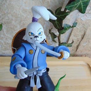 May include: A figurine of a white rabbit samurai with a blue kimono, black pants, and a straw hat. The rabbit holds a katana and a smaller knife. The figure has a stern expression and is posed as if ready for battle.