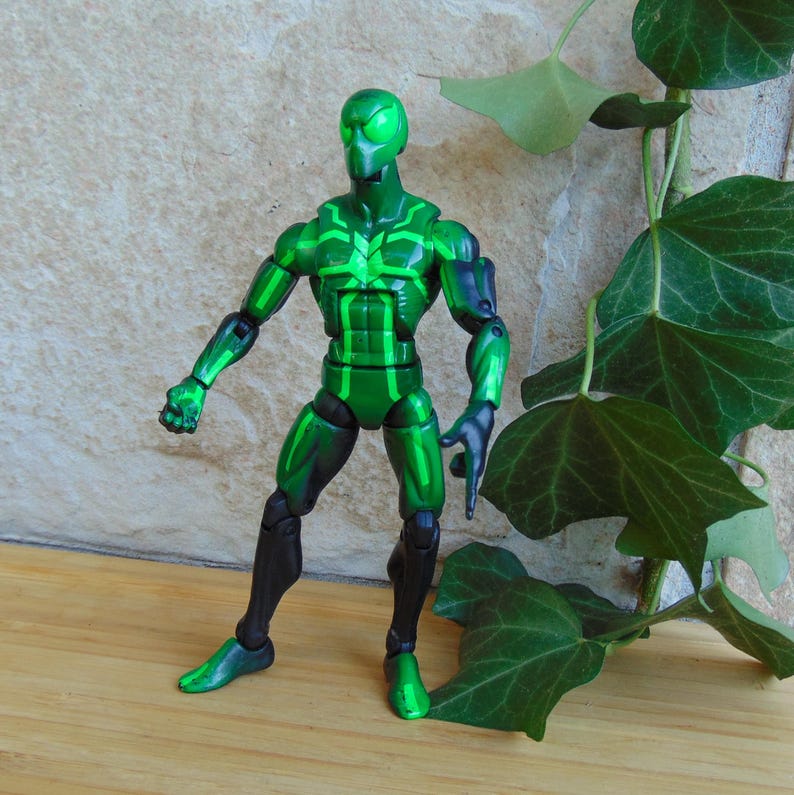 May include: A green action figure with black accents. The figure has a detailed design with a helmet and a muscular build. The figure is posed with one arm extended and the other bent. The figure is standing on a wooden surface.