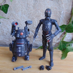 Könnte beinhalten: Zwei Star Wars-Actionfiguren: ein dunkelgrauer Droide mit roten Akzenten und ein humanoider Roboter mit silbernem Kopf und roten Details. Zubehör umfasst kleine schwarze Teile und ein silbernes Werkzeug. Die Figuren stehen auf einer Holzoberfläche.