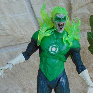 Peut inclure: Une figurine articulée Green Lantern avec un effet de flamme vert néon translucide émanant de la tête. La figurine porte une combinaison verte et bleu foncé avec un emblème circulaire central, des gants blancs et une expression faciale hargneuse.