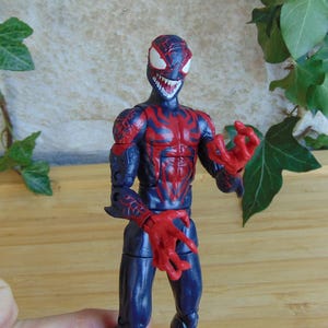 May include: A detailed action figure of a character in a dark blue and red color scheme. The figure has a menacing expression with sharp teeth and claw-like hands. The figure is posed with its hands outstretched.