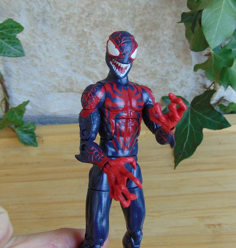 May include: A detailed action figure of a character in a dark blue and red color scheme. The figure has a menacing expression with sharp teeth and claw-like hands. The figure is posed with its hands outstretched.