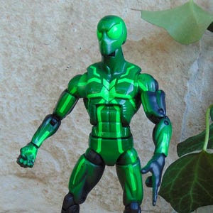 May include: A green action figure with a detailed design. The figure has a green head, torso, and limbs with lighter green accents. The figure is posed with one arm extended and the other bent. The figure is set against a textured background.