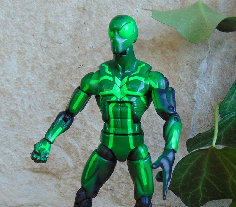 May include: A green action figure with a detailed design. The figure has a green head, torso, and limbs with lighter green accents. The figure is posed with one arm extended and the other bent. The figure is set against a textured background.