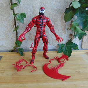 May include: A red and black action figure of a character with a white mask and claw-like hands. The figure is standing on a wooden surface, with red accessories scattered around it. Green leaves and vines are in the background.
