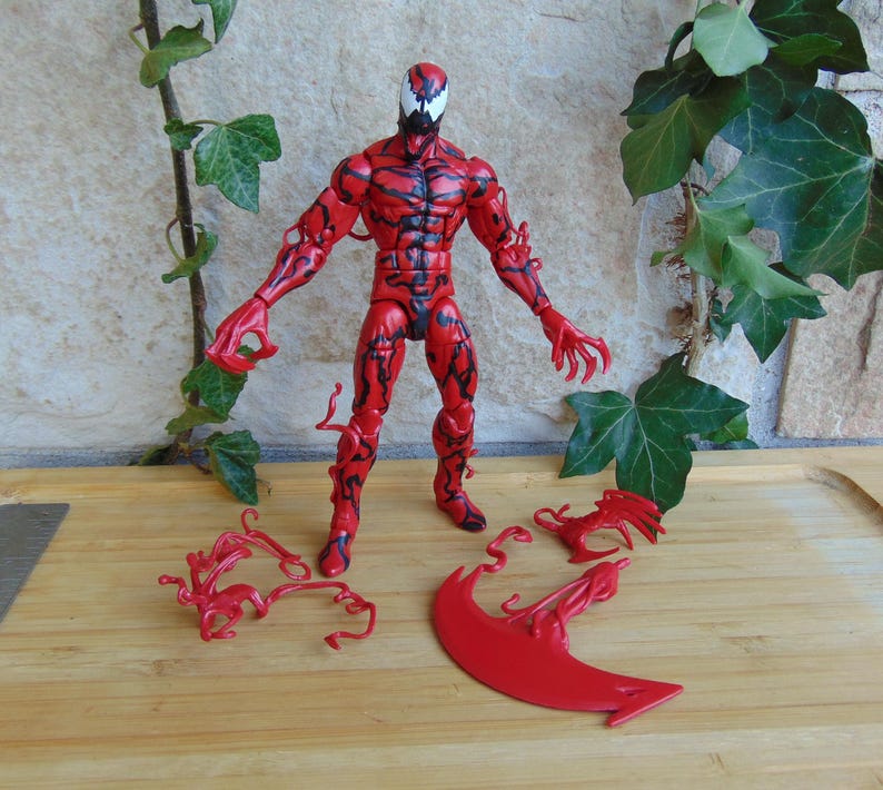 May include: A red and black action figure of a character with a white mask and claw-like hands. The figure is standing on a wooden surface, with red accessories scattered around it. Green leaves and vines are in the background.