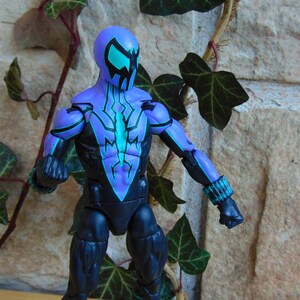 May include: A purple and black action figure with a teal and black spider design on the chest. The figure has a black mask with teal eyes.