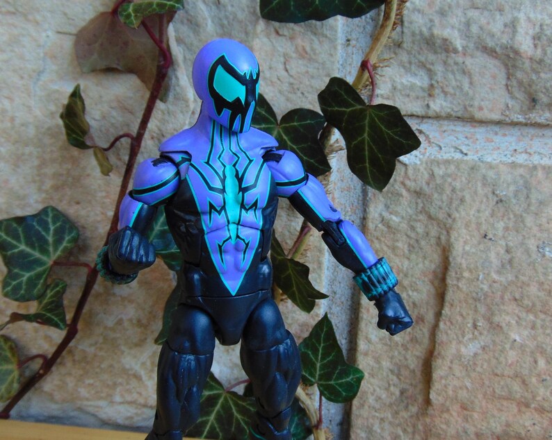 May include: A purple and black action figure with a teal and black spider design on the chest. The figure has a black mask with teal eyes.