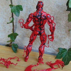May include: A red action figure of a character with black vein-like markings. The figure has multiple points of articulation and comes with detachable accessories. The figure is standing on a wooden surface with green ivy in the background.