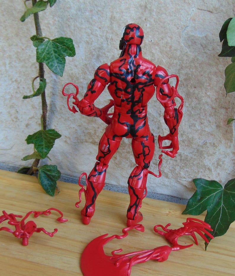 May include: A red action figure of a character with black vein-like markings. The figure has multiple points of articulation and comes with detachable accessories. The figure is standing on a wooden surface with green ivy in the background.