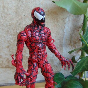 May include: A red Carnage action figure, a Marvel Comics character. The figure has a detailed, textured design with black veins and a white spider-like symbol on the head. The figure is posed with arms outstretched.