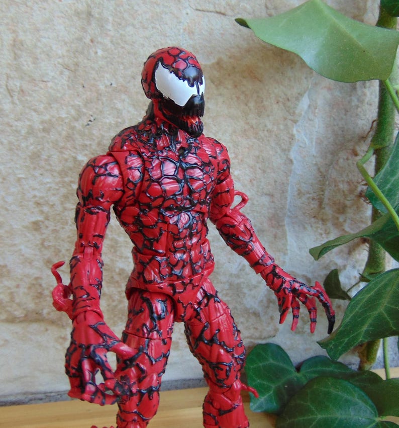 May include: A red Carnage action figure, a Marvel Comics character. The figure has a detailed, textured design with black veins and a white spider-like symbol on the head. The figure is posed with arms outstretched.