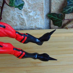 May include: Close-up of a red and black action figure's legs with pointed black boots. The figure is posed with one leg bent and the other leg straight out.