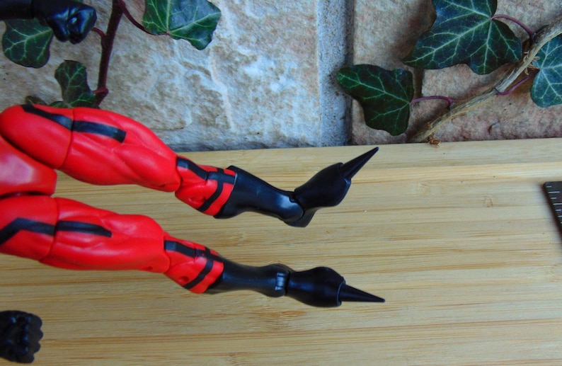 May include: Close-up of a red and black action figure's legs with pointed black boots. The figure is posed with one leg bent and the other leg straight out.
