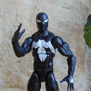 May include: A black action figure of a superhero with a white spider emblem on the chest. The figure has white eyes and a black mask. The figure is posed with one hand raised and the other at its side.