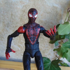 May include: A Miles Morales Spider-Man action figure. The figure is predominantly black with red accents, including a spider-web pattern on the mask and torso. The figure is posed with arms outstretched, showcasing the red hands.