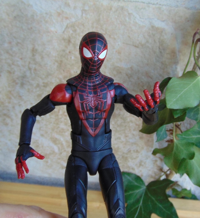May include: A Miles Morales Spider-Man action figure. The figure is predominantly black with red accents, including a spider-web pattern on the mask and torso. The figure is posed with arms outstretched, showcasing the red hands.