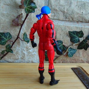 May include: A red and black action figure with a blue bandana on its head. The figure is posed with its arms at its sides and its legs slightly apart. The figure is standing on a wooden surface.
