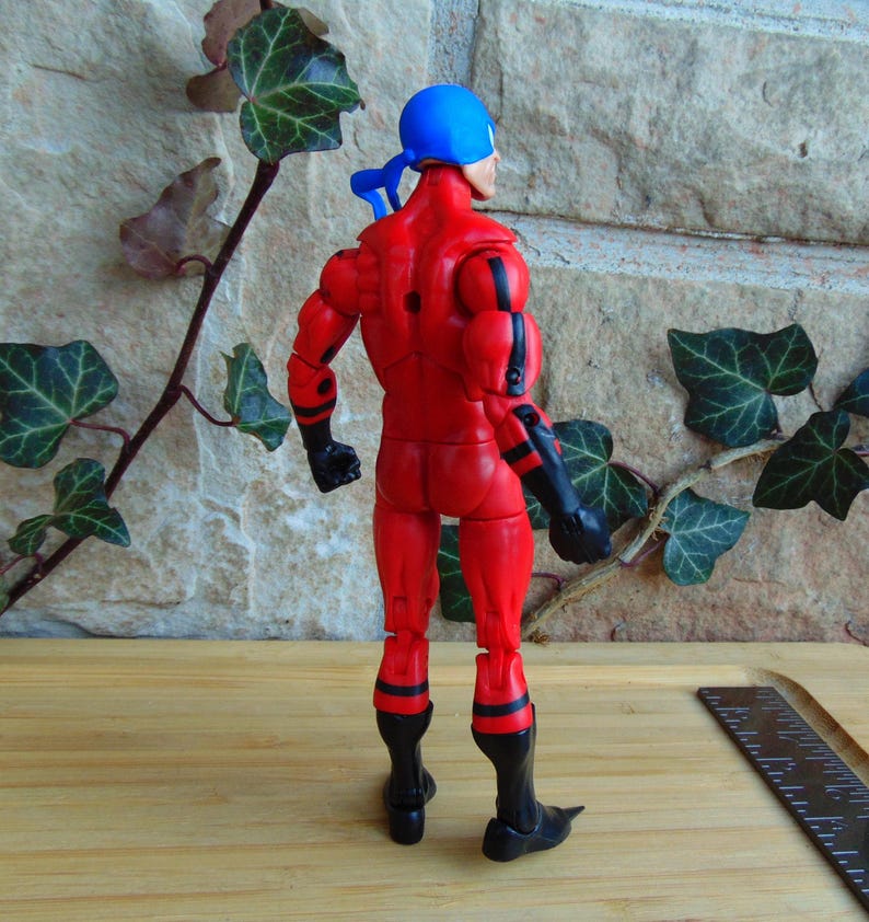 May include: A red and black action figure with a blue bandana on its head. The figure is posed with its arms at its sides and its legs slightly apart. The figure is standing on a wooden surface.