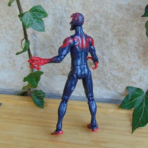 May include: A blue action figure with red accents, featuring a detailed design. The figure has red hands and feet, with red markings on the back and head. The figure is posed on a wooden surface, with green leaves in the background.