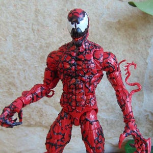 May include: A red action figure of the Marvel Comics character Carnage. The figure has a detailed, textured design with black veins and a white mask with black accents. The figure is posed with arms outstretched.