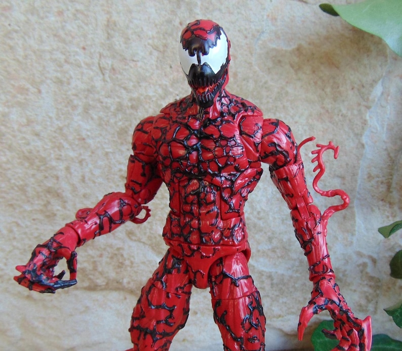 May include: A red action figure of the Marvel Comics character Carnage. The figure has a detailed, textured design with black veins and a white mask with black accents. The figure is posed with arms outstretched.