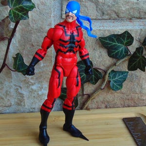 May include: Action figure of a superhero wearing a red and black suit with a blue mask and a black and red spider design on the chest. The figure has a mustache and is standing on a wooden surface.