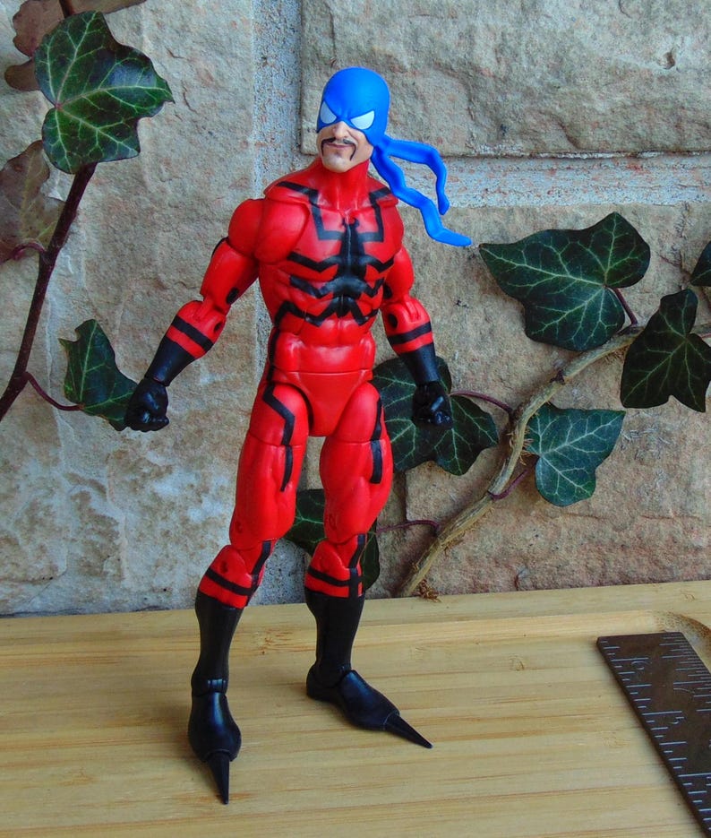 May include: Action figure of a superhero wearing a red and black suit with a blue mask and a black and red spider design on the chest. The figure has a mustache and is standing on a wooden surface.