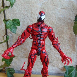 May include: A red action figure of the Marvel Comics character Carnage, with black vein-like markings. The figure has a white face with black accents and is posed with arms outstretched. The figure is approximately 6 inches tall.