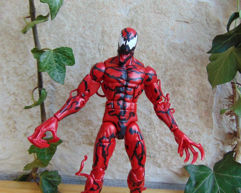 May include: A red action figure of the Marvel Comics character Carnage, with black vein-like markings. The figure has a white face with black accents and is posed with arms outstretched. The figure is approximately 6 inches tall.
